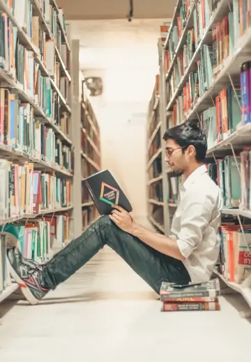 A man seated on the library floor, engrossed in reading a book amidst shelves filled with various titles