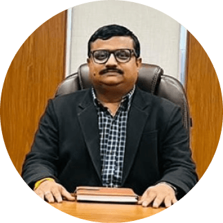 A professional man in a black suit and glasses sits at a desk with his hands clasped over a closed book, smiling slightly, in a well-appointed office, representing the successful guidance provided by Tajhind