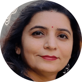 Close-up of a South Asian woman with shoulder-length dark hair, wearing a gray top and small earrings, featuring a subtle smile against a blurred background. She has a red bindi on her forehead and