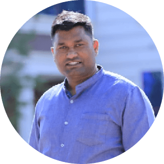 A smiling man in a blue shirt standing outdoors, with a blurred background suggesting a sunny, urban setting near Tajhind Edutech Pvt Ltd.