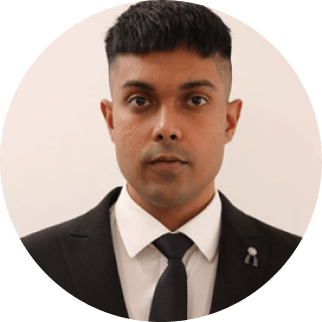 A portrait of a man in a black suit and tie, with styled hair, looking directly at the camera against a plain light background. he wears a lapel pin on his left side.