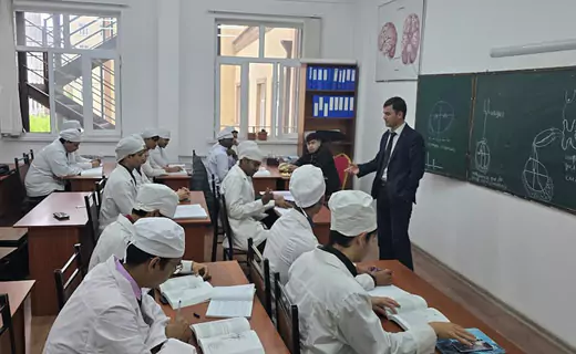 A classroom with a group of students in white lab coats taking notes while a teacher in a dark suit lectures and points to a chalkboard with anatomical drawings at TajHind Edutech Pvt Ltd.