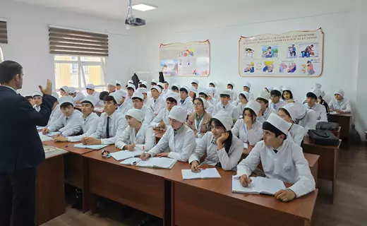 A classroom scene with a teacher lecturing to students wearing white uniforms and caps, representing an MBBS study abroad consultancy. The room has educational posters on the walls, and students are seated at desks with